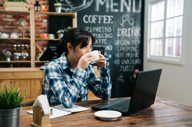 Asiatische elegante freiberufliche frau, die kaffee trinkt und eine heiße tasse in der vintage-café-bar hält, während sie an einem laptop-computer arbeitet, der eine pause macht. junges Mädchen Kleinunternehmer mit Blick aus dem Fenster denken