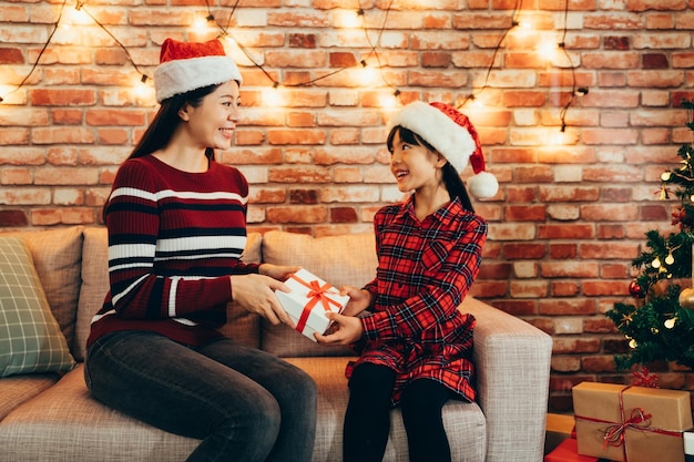 asiatische familie feiert zu hause am heiligabend. Zwei Mädchen mit roten Hüten tauschen Weihnachtsgeschenke aus und sitzen auf einer bequemen Couch in einem dekorierten Haus. mutter und kind starke fröhliche liebe.