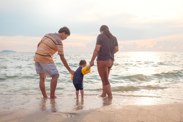 Asiatische Familie mit 2 Jahren alten Kleinkindbabykind barfuß gehend auf den Strand im Wasser bei Sonnenuntergang.