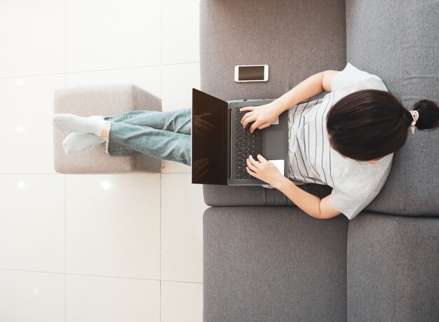 Asiatische Frau arbeiten zu Hause mit Notebook zur Konferenz mit Online-Team, sitzen bequem auf dem Sofa im Wohnzimmer, arbeiten von zu Hause aus.