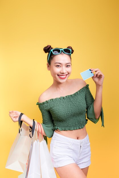 Asiatische Frau beim Einkaufen hält Einkaufstüten und eine Kreditkarte auf gelbem Hintergrund