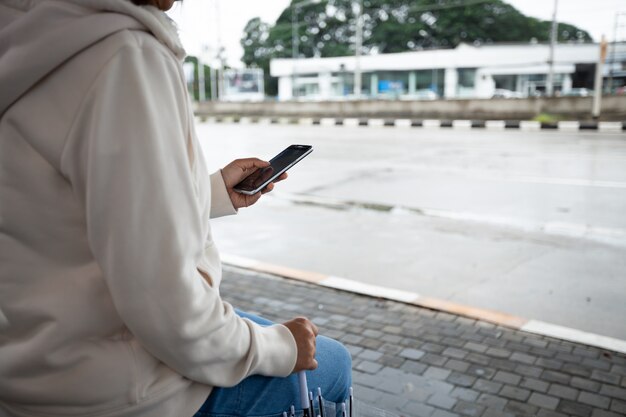 Asiatische Frau benutzt auf dem Smartphone und überprüft das soziale Mediennetzwerk, während sie am regnerischen Tag an der Bushaltestelle auf ein Taxi wartet.