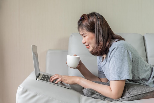 Asiatische Frau benutzt Computer-Notebook und trinkt Kaffee auf dem Sofa