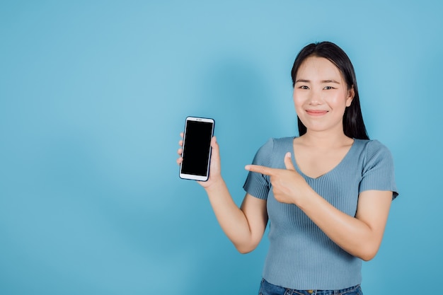 Asiatische Frau blaues stehendes Abnutzungshemd, das mit dem Finger auf den leeren Bildschirm des intelligenten digitalen Handys auf blauem Hintergrund zeigt