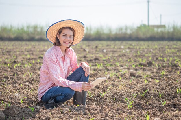 Asiatische Frau des jungen Bauern, die intelligente Tablette in einem Maisfeld sitzt und hält. Technologiefarm Konzeptbild.