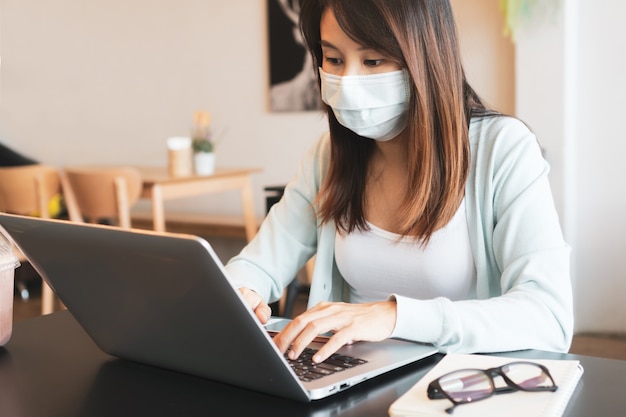 asiatische Frau, die am Computer arbeitet, während sie während der Coronavirus-Krise eine medizinische Maske im Café trägt