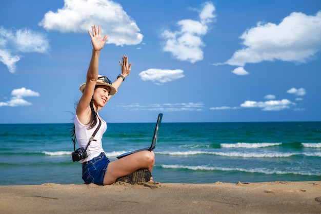 Asiatische Frau, die am Strand sitzt und Laptop für ihre Arbeit benutzt und Geschäft während ihres Urlaubs prüft.