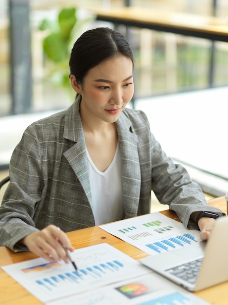 Asiatische Frau, die an Laptop arbeitet. Geschäftsfrau beschäftigt, an ihrem Finanzprojekt zu arbeiten. Finanzberichte auf dem Tisch