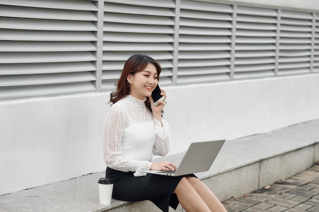 Asiatische Frau, die auf dem Handy spricht, während sie einen Computerlaptop benutzt