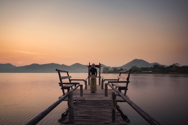 Asiatische Frau, die auf dem Pier steht und den Sonnenaufgang auf dem Berg am Stausee betrachtet