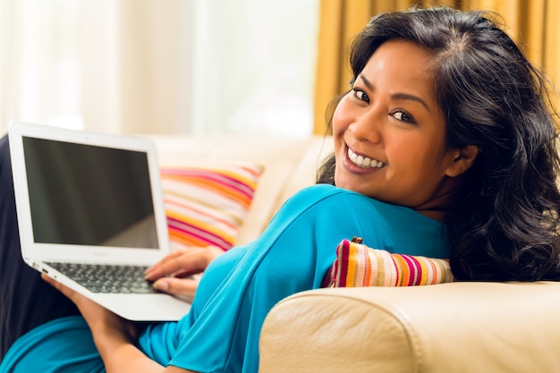 Asiatische Frau, die auf der Couch surft das Internet und lächelt sitzt