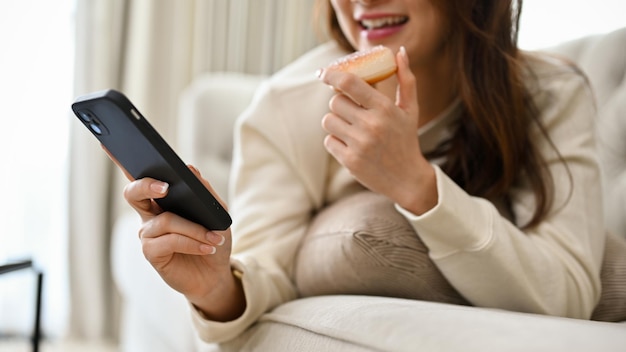 Asiatische Frau, die auf einem bequemen Sofa liegt, Donut isst und ein beschnittenes Bild des Smartphones verwendet