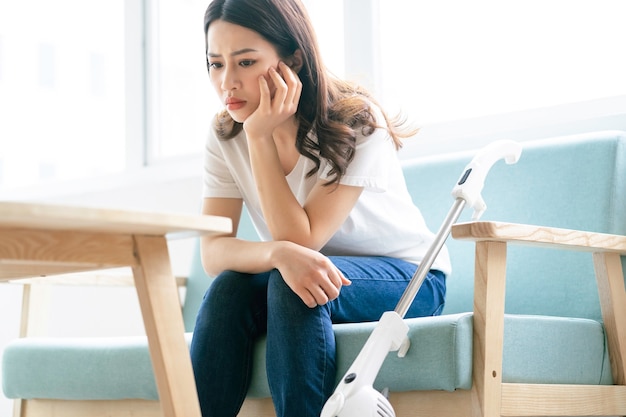 Asiatische Frau, die auf einem Stuhl mit einem müden Ausdruck beim Reinigen des Hauses sitzt