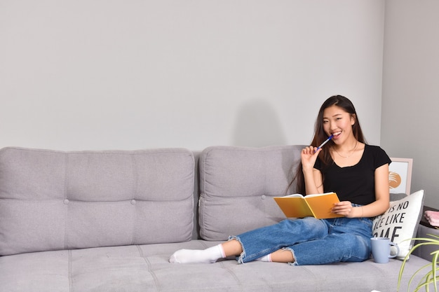 Asiatische Frau, die auf Sofa und orangeem Heft sitzt