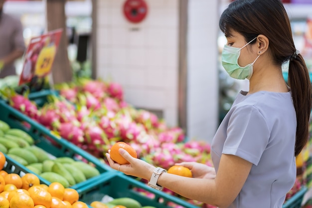 Asiatische Frau, die chirurgische Gesichtsmaske trägt und Orangenfrucht im Supermarkt hält