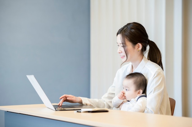 Asiatische Frau, die ein Baby hält und einen Laptop drinnen bedient