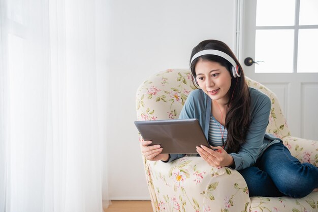 asiatische frau, die ein tablet hält und einen kopfhörer trägt, während sie auf der couch im wohnzimmer sitzt.