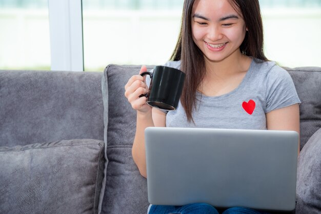 Asiatische Frau, die eine Kaffeetasse lächelt und während einer Arbeit schaut, aus der Ferne mit einem Laptop auf dem Sofa im Haus arbeitet, süße Mädchen-Teenager verwenden gerne Internet-Computer, um von zu Hause aus zu kommunizieren