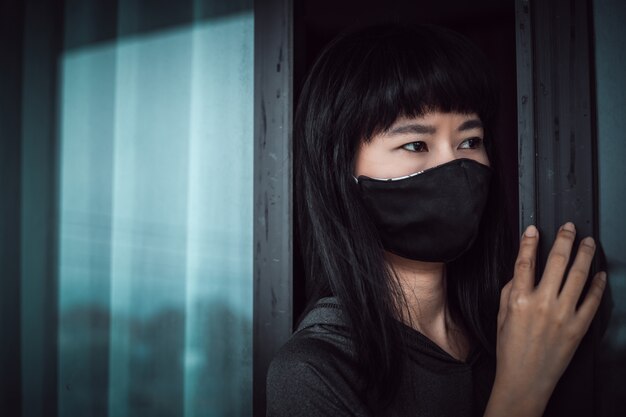 Asiatische frau, die eine schwarze gesichtsmaske trägt, geistesabwesend an der tür steht und zu hause für selbstquarantäne und soziale distanzierung im coronavirus- oder covid-2019-ausbruchssituationskonzept bleibt