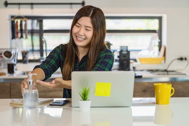 Asiatische Frau, die Händedesinfektionsmittel verwendet, wenn sie zu Hause mit einem Technologie-Laptop arbeitet