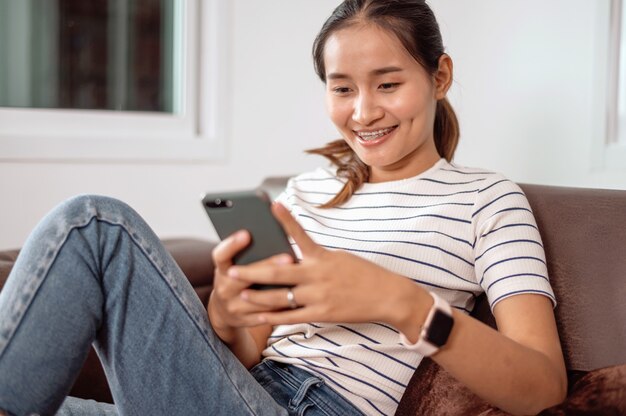 Asiatische Frau, die Handy auf Sofa benutzt und zu Hause lächelt