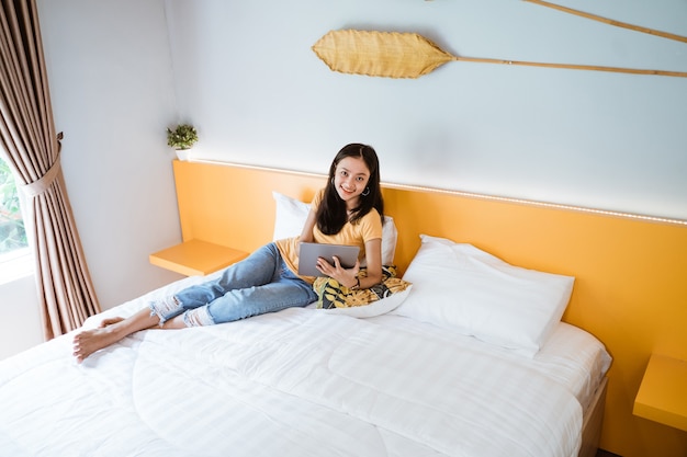 Asiatische Frau, die im Bett mit digitaler Tablette liegt, die mit Finger im Morgensonnenlicht berührt