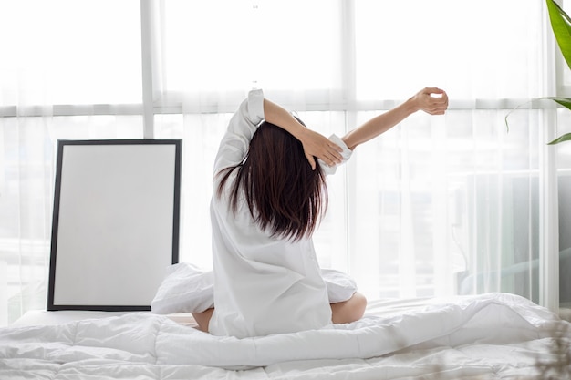 Asiatische Frau, die im weißen Bett sitzt und schläft und sich am Morgen im Schlafzimmer ausdehnt