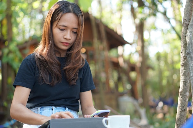 Asiatische Frau, die in einem Café, in dem sie inmitten grüner Natur zur Arbeit gehen kann, auf ihrem Telefon die Einnahmen ansieht