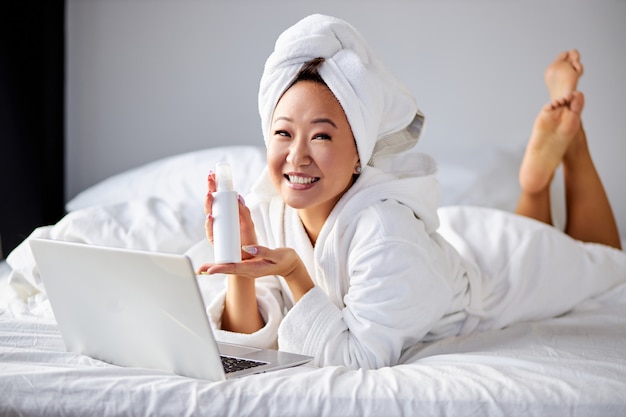 Asiatische Frau, die Komsetik-Sahneflasche in den Händen hält, mit Laptop auf Bett liegend, sie bewirbt das Produkt