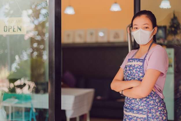 Asiatische Frau, die Maske steht mit offenem Schild am Fenster im Kaffeehaus steht.