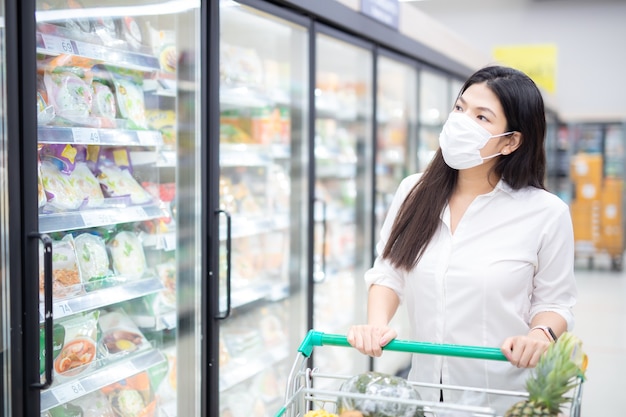 Asiatische Frau, die mit Maske einkauft, die sicher für Lebensmittel, Sicherheitsmaßnahmen im Supermarkt kauft.