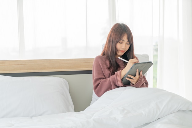 Asiatische Frau, die mit Tablette auf Bett arbeitet