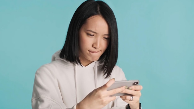 Asiatische Frau, die nervös aussieht und emotional auf dem Smartphone im Online-Spiel auf blauem Hintergrund spielt