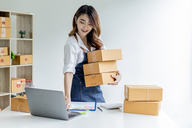 Asiatische Frau, die Paketkästen hält und Bestellungen vom Laptop aus überprüft, sie besitzt einen Online-Shop, sie verpackt und versendet über ein privates Transportunternehmen. Online-Verkaufs- und Online-Shopping-Konzepte.