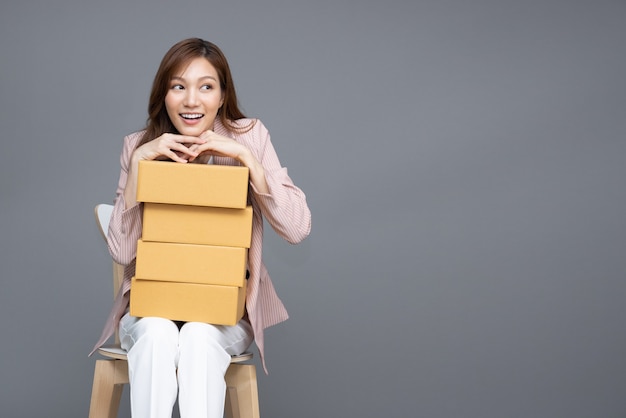 Asiatische Frau, die Paketkasten hält und auf weißem Stuhl sitzt, isoliert auf grauem Hintergrund