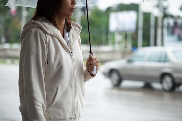 Asiatische Frau, die Regenschirm hält, während Taxi wartet und auf der Bürgersteigstraße der Stadt im regnerischen Tag steht.