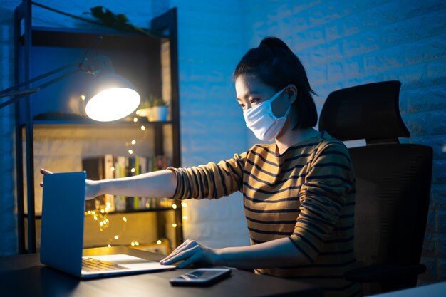 Asiatische Frau, die Schutzgesichtsmaske trägt, die an einem Laptop Überstunden zu Hause arbeitet. . Arbeiten Sie von zu Hause aus, um das Coronavirus COVID 19-Konzept zu vermeiden.