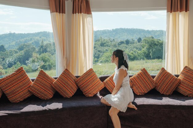 Asiatische Frau, die sich entspannt und den Blick auf den tropischen Wald auf dem Sofa im Panoramazimmer am hellen Tag genießt