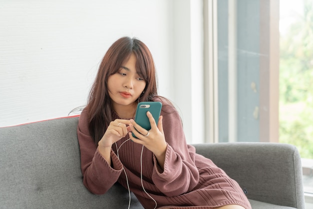 Asiatische Frau, die Smartphone auf grauem Sofa im Wohnzimmer verwendet