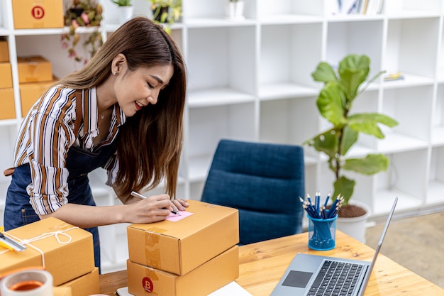 Asiatische Frau, die Versandinformationen des Kunden auf Paketkasten schreibt, sie besitzt einen Online-Shop, sie versendet Produkte über ein privates Kurierunternehmen an Kunden. Konzept für den Online-Verkauf.