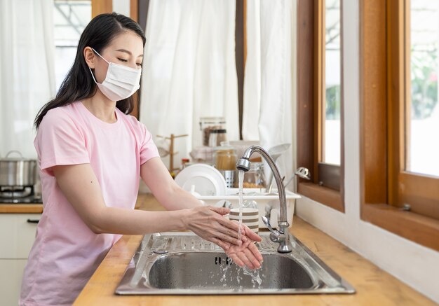 Foto asiatische frau, die von zu hause aus arbeitet, schutz- oder gesichtsmaske tragend und ihre hände mit sauberem wasser in der küche zu hause während der coronavirus-krise oder des ausbruchs der covid-19 wäschend.