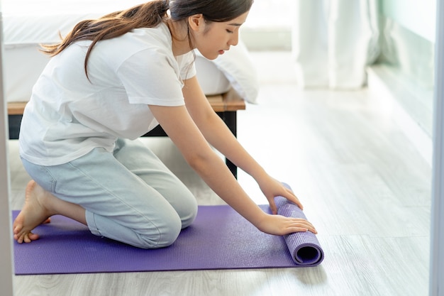 Asiatische Frau, die Yogamatte ausbreitet, um Yoga zu Hause vorzubereiten