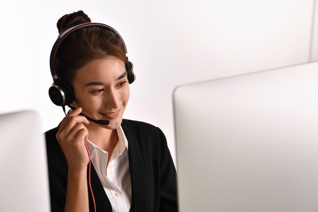 Asiatische Frau im Callcenter, die ein Headset verwendet, schaut auf den Computerbildschirm. Porträt einer lächelnden asiatischen Kundendienstmitarbeiterin mit Headset, die im Hintergrund im Büro arbeitet