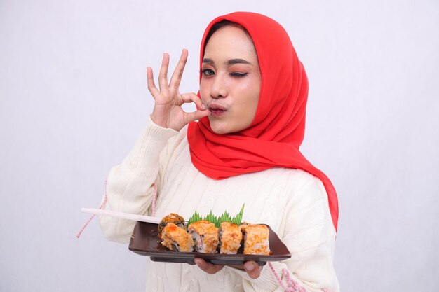 Asiatische Frau im Hijab, die sich küsst, zwinkt mit einer köstlichen Geste in die Kamera und beide Hände halten eine Platte