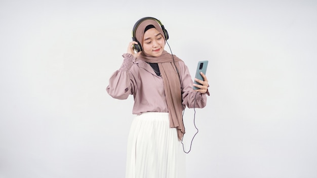Asiatische Frau im Hijab, die zuhört, während sie auf ihren Telefonbildschirm schaut, der auf weißem Hintergrund isoliert ist