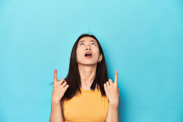 Asiatische Frau im sommergelben Top-Studio-Setup zeigt nach oben mit geöffnetem Mund