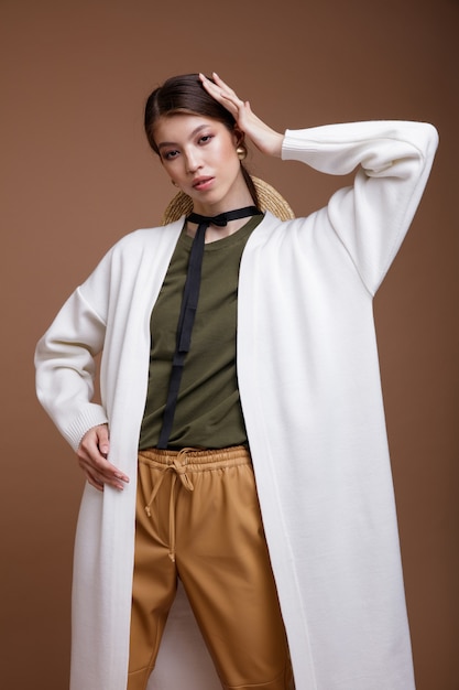 Asiatische Frau in beige Lederhose weiße lange Strickjacke grüner T-Shirt Gürtel auf braunem Hintergrund