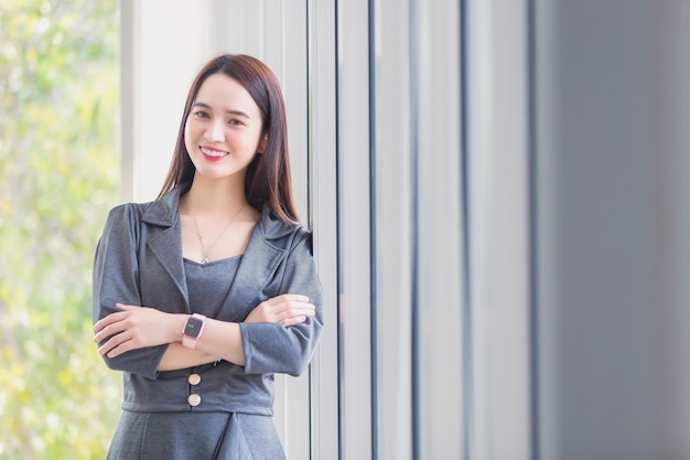 Asiatische Frau in dunkelgrauem Kleid lächelt glücklich und steht neben grauem Vorhang am Arbeitsplatz.