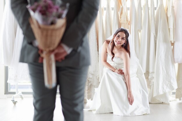 Asiatische Frau in einem Brautkleid Lächelnder Bräutigam. Der Bräutigam im grauen Anzug hält einen lila Blumenstrauß, um die Braut zu überraschen, die in der Umkleidekabine sitzt. Konzept Hochzeit besten Tag.