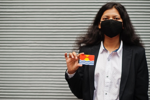 Asiatische frau in formeller kleidung und schwarzer gesichtsmaske halten kiribati-flagge vor grauem hintergrund zur hand coronavirus im länderkonzept